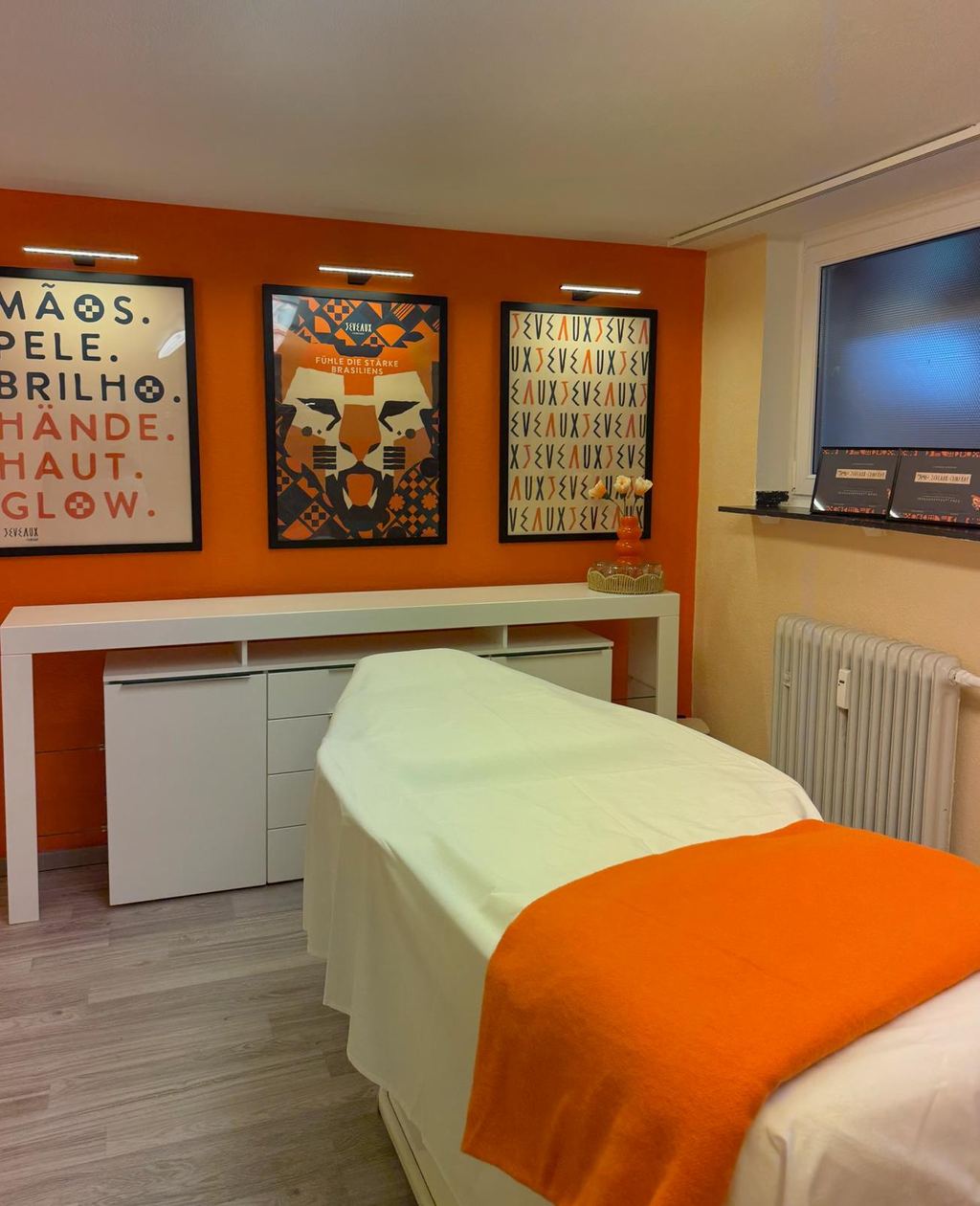 Behandlungsraum bei Shape and Flow: Massageliege mit orangefarbenem Plaid vor der orangefarbenen Wand mit Jeveaux-Postern